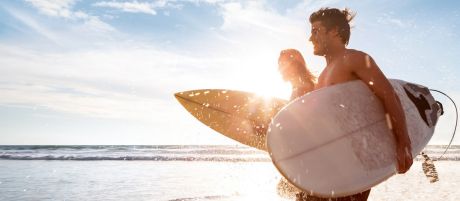 Ein junges Pärchen ist mit Ihren Surfbrettern unter den Armen auf dem Weg ins Meer und freut sich über einen schönen sonnigen Tag